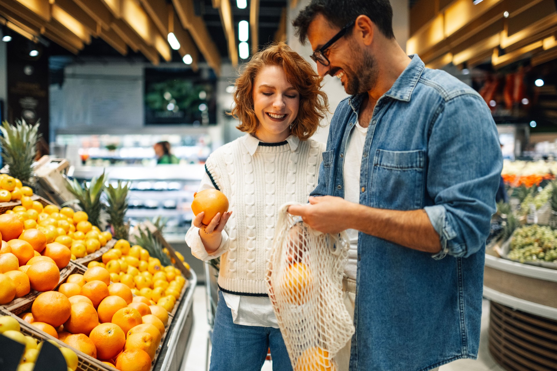 Happy couple choosing oranges in reusable mesh bag at grocery store