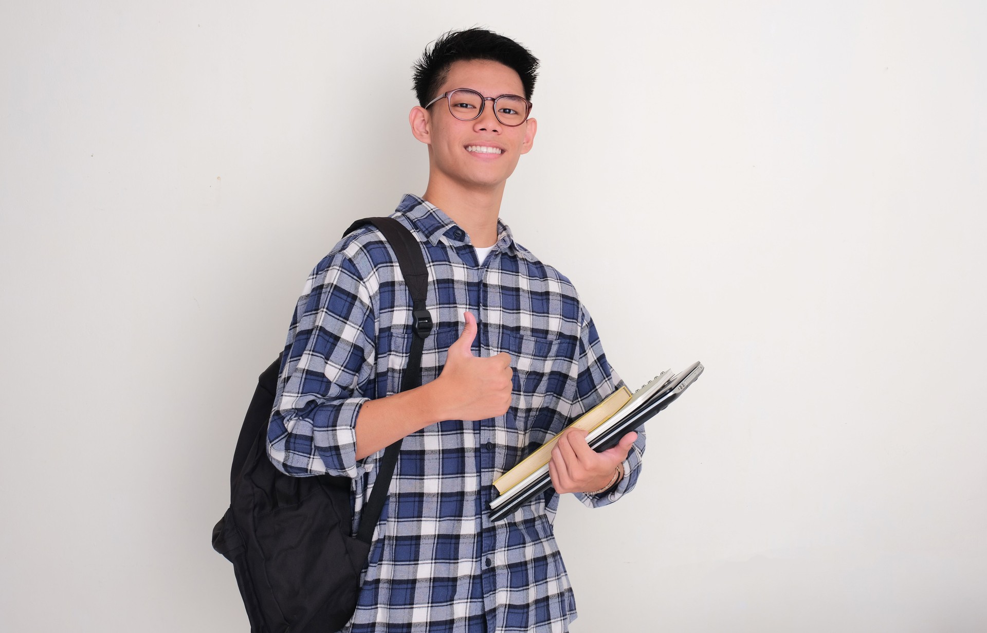 A male college student smiling and give thumbs up while holding books, laptop and wearing backpack