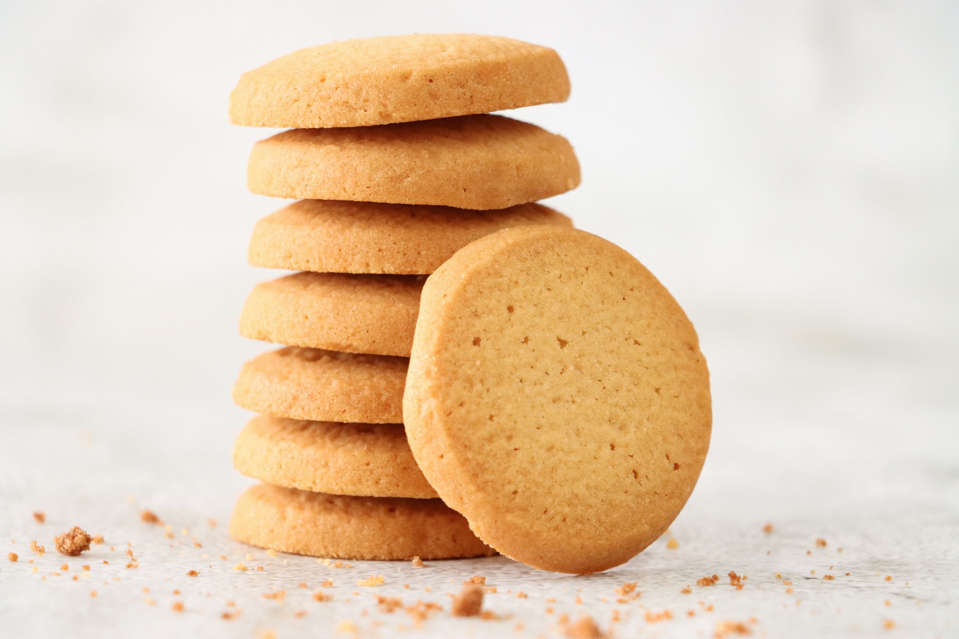 Close-up image of shortbread round leaning against stack of freshly baked, homemade shortcake biscuits surrounded by crumbs, mottled white background, focus on foreground