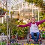 Photo of young Paralyzed young man, sitting and looking at camera with raised his hands on his wheel chair against greenhouse during the day. Front view of a Young male gardener working in the greenhouse with arms up